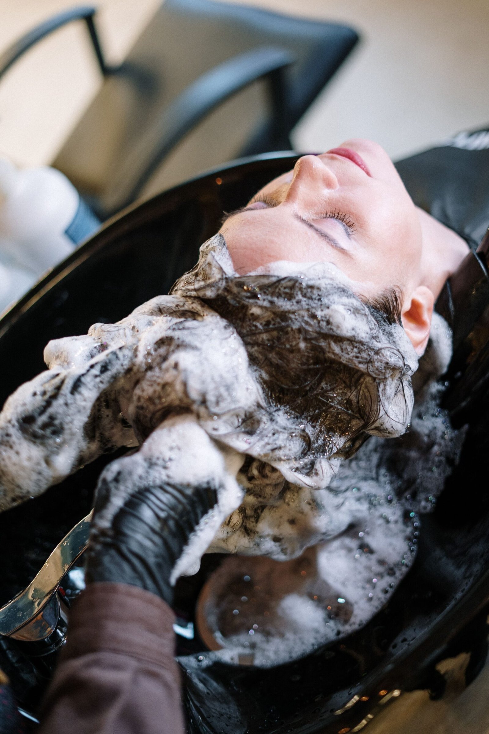 Woman receiving a professional shampoo treatment at a salon sink, with her hair covered in cleansing foam.